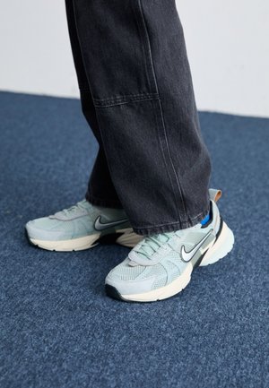 Világoskék hálós Nike sportcipők texturált felülettel, fekete swoosh-szal és krémszínű talprésszel, sötétszürke laza nadrággal párosítva.