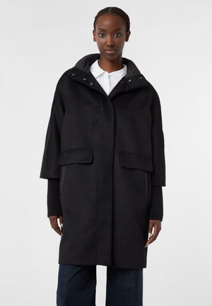 Cappotto di lana nera con cappuccio, bottoni a pressione frontali, due tasche anteriori e polsini a coste. Texture morbida con una forma strutturata e design fino al ginocchio.