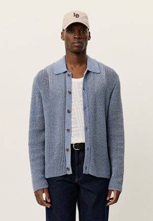 Lyseblå strikket cardigan med krave, knaplukning foran og struktureret mønster. Båret over en hvid skjorte og mørke denimbukser.