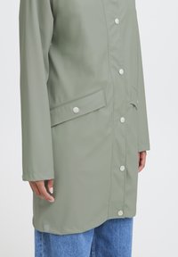 Giacca impermeabile verde chiaro con bottoni a pressione e una tasca laterale, caratterizzata da una linea dritta e una texture liscia. Indossata sopra un jeans blu.