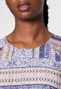 Top multicolor com padrão, apresentando um decote redondo. O design inclui padrões geométricos intrincados em tons de azul, bege e marinho.