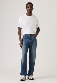 Άνδρας με λευκό κοντομάνικο μπλουζάκι, συνδυασμένο με μπλε τζιν παντελόνι ίσιας γραμμής με ξεβαμμένο εφέ, και αθλητικά παπούτσια σε ανοιχτό γκρι χρώμα.