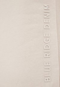 Svetlo bež tkanina z teksturirano površino, ki vsebuje dvignjene bele črke: "BLUE RIDGE DENIM." Preprost dizajn, minimalni akcenti.