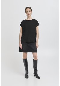 Naine, kes seisab musta lühikeste varrukatega pluusi, musta seelikuga ja põlvikuni musta nahast saapaga ühetaolise heleda tausta ees.