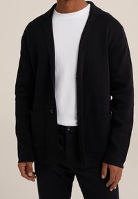 Svart stickad cardigan med skarpt slag, öppen front och två framfickor. Bärs över en vit t-shirt med rund halsringning.