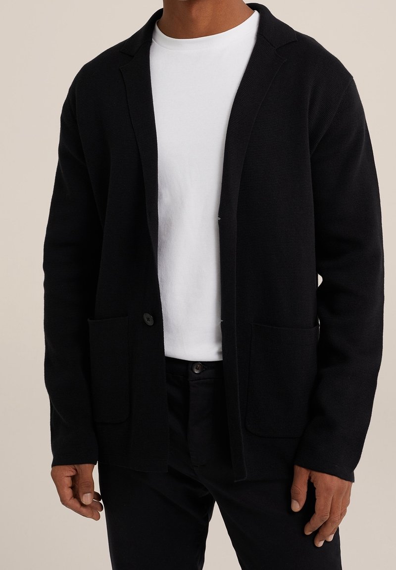 Svart stickad cardigan med skarpt slag, öppen front och två framfickor. Bärs över en vit t-shirt med rund halsringning.