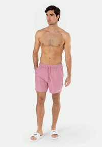 Ροζ μαγιό με ελαστική μέση και κορδόνι. Υφασμάτινη υφή; το μοντέλο φοράει λευκά σανδάλια. Το φόντο είναι απλό λευκό.