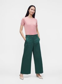 Femme aux longs cheveux foncés portant un haut rose à manches courtes, un pantalon vert large et des chaussures beiges pointues, debout les mains dans les poches.