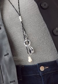 Schwarze Lederjacke über einem grau gestreiften Pullover, mit einer silbernen Anhängerhalskette mit ovalen und tropfenförmigen Elementen und einem weißen Perlakzent.