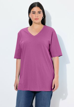 Frau mit dunklen Haaren trägt ein übergroßes lila T-Shirt mit V-Ausschnitt und blaue Jeans, steht vor einem weißen Hintergrund.