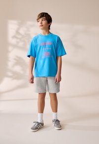 Tricou albastru din bumbac cu text grafic roz, asortat cu pantaloni scurți gri. Modelul poartă șosete albe și adidași gri cu accente negre.