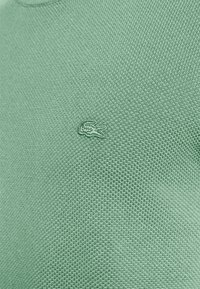 Grüner gestrickter Pullover mit einem strukturierten Muster, versehen mit einem kleinen, gestickten Logo eines Krokodils auf der linken Brustseite.