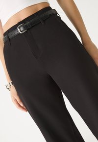 Schwarze maßgeschneiderte Hose mit einer glatten Textur, ausgestattet mit einem Bund und einem schwarzen Gürtel mit silberner Schnalle. Seitentaschen sind sichtbar.