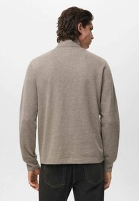 Uomo con capelli ondulati scuri che indossa un maglione di maglia beige a maniche lunghe e jeans neri, di spalle su uno sfondo chiaro.