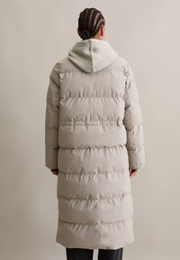Lang, gewatteerde jas in lichtbeige met een geribbelde grijze hoodie. Heeft doorgestikte secties en een rechte snit. Zachte, gladde textuur.