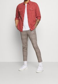 Rood overhemd met knopen, witte T-shirt eronder, geruite broek in bruin en beige, witte sneakers, staand tegen een lichtgrijze achtergrond.