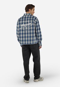 Homme portant une chemise à carreaux bleue et blanche avec un texte blanc dans le dos, pantalon noir et baskets grises, debout de dos.