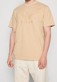 Camiseta de algodón beige con mangas cortas, que presenta un logo de Adidas texturizado y tonal, junto con un gráfico de hojas en la parte frontal.