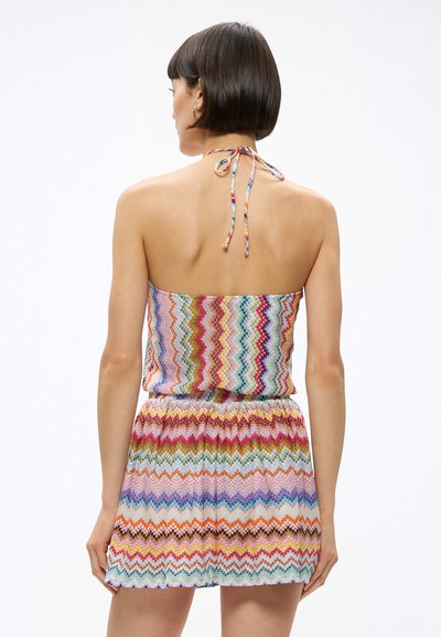 Γυναίκα με κοντά σκούρα μαλλιά που φοράει πολύχρωμο φόρεμα με σχέδιο ζιγκ-ζαγκ και λαιμόκοψη halter, γυρισμένη με την πλάτη προς τα έξω, σε απλό λευκό φόντο.