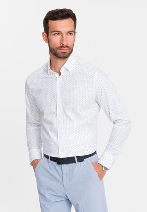 Camicia bianca con piccoli pois neri, colletto button-down, maniche lunghe e vestibilità slim, abbinata a pantaloni azzurri chiari e cintura nera.