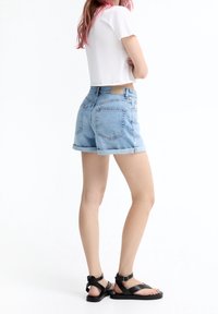 Shorts de mezclilla azul claro con dobladillos enrollados, con bolsillos traseros y una etiqueta de marca marrón. Combinados con sandalias de cuero negras.