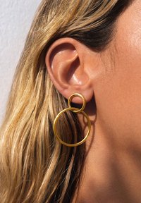 Pendiente de aro dorado con un diseño circular doble, que presenta una textura suave. Se muestra parcialmente el cabello y una oreja en el encuadre.