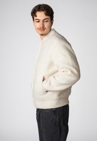 Cream-colored fleece bomber jacket with a ribbed collar and cuffs, featuring side pockets and a relaxed fit. Worn with dark jeans.