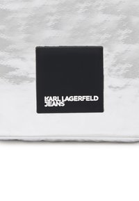 Materiale bianco testurizzato con un'etichetta quadrata nera che presenta la scritta "KARL LAGERFELD JEANS" in testo argentato. Design semplice e moderno.