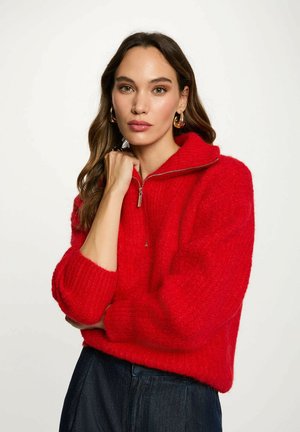 Maglione rosso in maglia soffice con colletto con zip, polsini a coste e vestibilità rilassata, abbinato a denim scuro. Accentuato da orecchini a cerchio dorati.