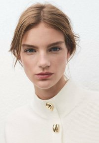 Jeune femme aux cheveux châtain clair et aux yeux bleus portant un haut blanc texturé avec des boutons dorés, regardant directement l'objectif.