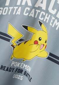Šedé tričko s grafickým znakom žltého Pikachu, text „POKEMON“ hore a „READY FOR BATTLE 0025“ dole, s čiernymi pruhmi ako akcentmi.