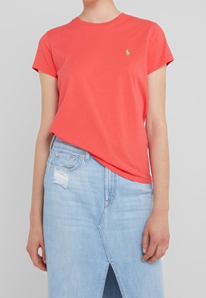 Osoba ubrana w koralowy t-shirt z krótkim rękawem z małym zielonym logo oraz jasnoniebieską spódnicę jeansową z przednim rozcięciem i przetartym naszywką.