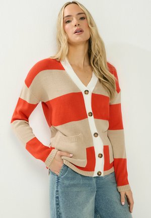 Frau trägt eine beige-rot gestreifte Strickjacke mit Holzknöpfen und hellblaue Jeans, steht vor einer weißen Wand.