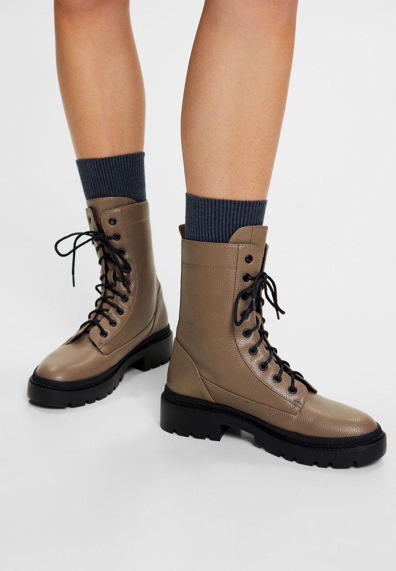 Personne portant des bottes de combat en cuir taupe à lacets avec des semelles épaisses noires et des chaussettes côtelées bleu foncé sur un fond uni.