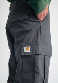 Pantalons cargo gris en tissu léger, dotés d'une poche latérale avec un logo Carhartt visible et une texture en grille.
