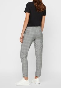 Kvinna i grå rutan slim-fit byxor, svart kortärmad topp och vita sneakers, stående mot en ljus, enfärgad bakgrund.