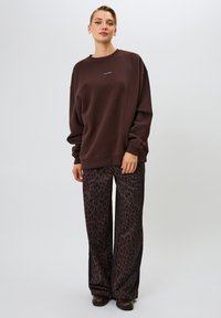 Hnedý oversized sveter s rebrovanými manžetami, skombinovaný s nohavicami širokého strihu s čiernym leopardím vzorom na tmavom pozadí.