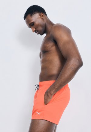 Άντρας χωρίς πουκάμισο με τα χέρια στις τσέπες, φοράει φωτεινά πορτοκαλί σορτς Puma, στέκεται πλαγίως μπροστά σε απλό ανοιχτό φόντο.