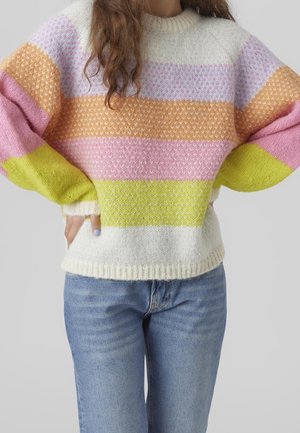 Osoba ubrana w wielokolorowy sweter w paski w kolorach białym, żółtym, różowym, pomarańczowym i lawendowym, w połączeniu z niebieskimi dżinsami i rękami na biodrach.