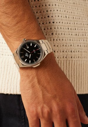 Silver metal wristwatch with black dial and date display worn on left wrist over knitted beige sweater and dark pants.