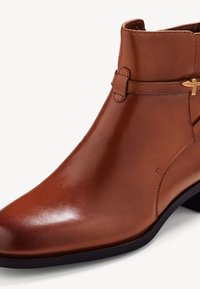 Bottine en cuir marron avec une finition lisse, bout pointu, petit talon, et une sangle décorative avec un accent doré.