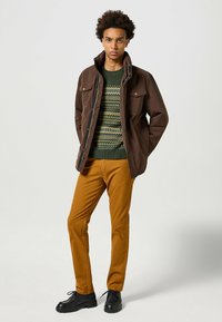 Brązowa kurtka z dwiema kieszeniami na piersi, zielony sweter w wzory i pomarańczowe spodnie. Czarne buty dopełniają stylizację. Gładka tekstura materiału.