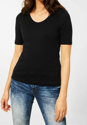 Kvinde iført en sort kortærmet V-hals t-shirt og blå falmede jeans, der står foran en ensfarvet baggrund.