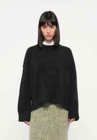 Svart oversized genser med rund hals, ribbestrikket kant og senkede skuldre, laget av et mykt, teksturert materiale. Kombinert med et lyst skjørt.