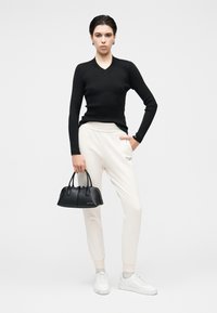 Top preto de manga comprida com textura canelada, calças de treino creme, ténis brancos e uma pequena mala preta. Modelo em pé sobre um fundo neutro.