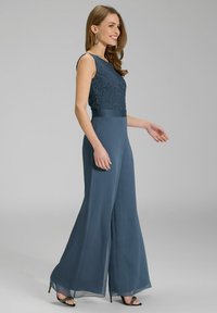 Combinaison sans manches avec un corsage en dentelle et un pantalon large et fluide. Le matériau est transparent et texturé, d'une couleur bleu profond. Ceinture d'accent à la taille.