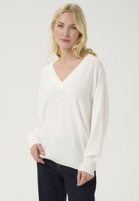 Balta V-veida džemperis, izgatavots no mīksta auduma, ar garām piedurknēm un ribotām aprocēm un apakšmalu, uzvilkts pār tumšajiem džinsiem.