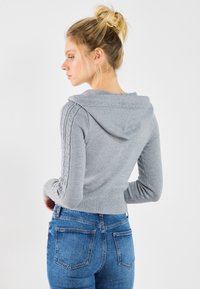 Gray cropped hoodie made of soft knit fabric, featuring a hood and cable knit patterns along the sleeves. Paired with blue jeans.