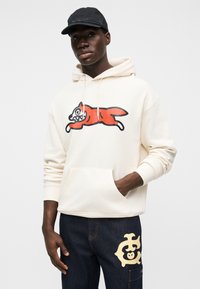 Κρεμ φούτερ με κουκούλα και φωτεινό κόκκινο γραφικό καρτούν αλεπούς, κορδόνι στην κουκούλα και μπροστινή τσέπη· συνδυασμένο με σκουρόχρωμο τζιν παντελόνι που διαθέτει μπεζ λογότυπο.