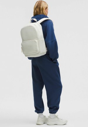 Zaino bianco con una superficie liscia, forma rettangolare, tasca frontale e spallacci regolabili, indossato sopra una tuta da ginnastica blu navy.